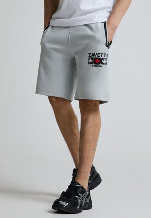Mand iført lysegrå ZAVETTI Canada shorts med sorte lynlåslommer og sorte løbesko står med hænderne i lommerne.