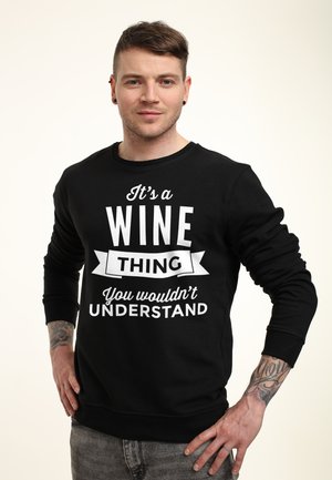 Man met tatoeages die een zwarte trui draagt met de tekst "It's a WINE THING You wouldn't UNDERSTAND", staand met handen in de zij.