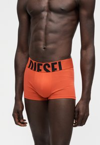 Torso masculin portant un boxer orange ajusté avec une large ceinture noire affichant le nom de la marque "DIESEL" en lettres grasses.