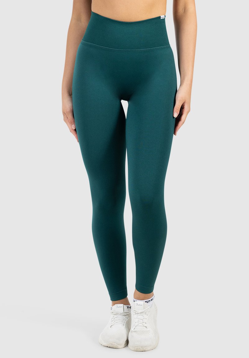 Femme portant un legging vert foncé taille haute et des chaussures de sport blanches, debout devant un fond clair uni.