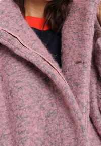 Personne portant un manteau en laine rose texturé par-dessus un haut col rond noir et rouge, mettant l'accent sur le col et la partie supérieure de la poitrine.