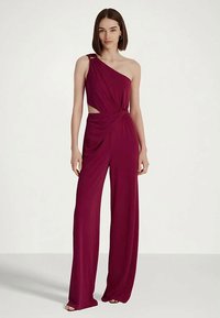 Egy nő ujjatlan, egyvállas, szilva színű, széles lábú kezeslábasban áll, amelynek oldalt kivágása van, egy sima világos háttér előtt.