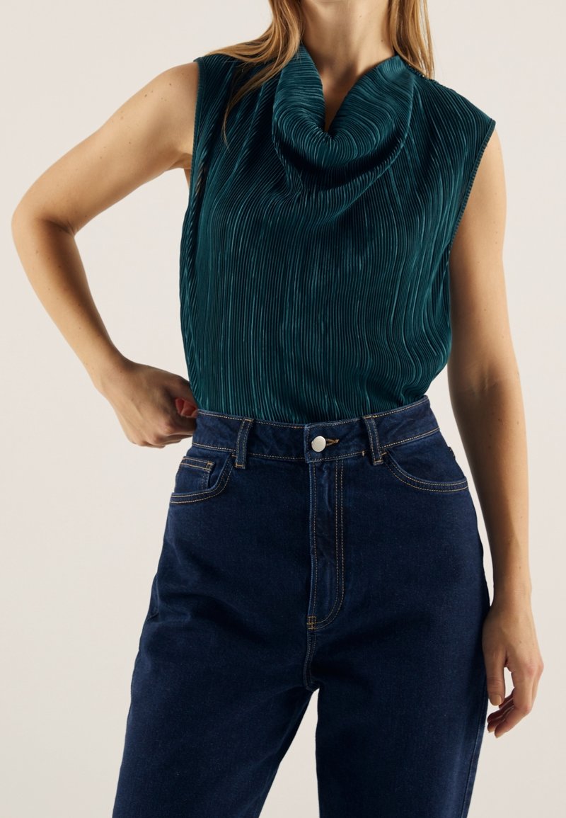 Femme portant un haut sans manches plissé bleu sarcelle foncé à col bénitier, rentré dans un jean taille haute bleu foncé.