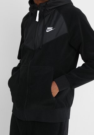 Sweat à capuche noir zippé Nike avec poches kangourou, capuche avec cordon et logo Nike blanc sur la poitrine, porté les mains dans les poches.