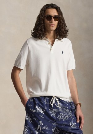 Νέος άνδρας με μακριά σγουρά μαλλιά που φοράει γυαλιά ηλίου, λευκό πόλο και μπλε σορτς με σχέδια, με τα χέρια στις τσέπες.