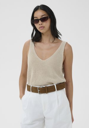 Donna con occhiali da sole scuri, top beige senza maniche in maglia, pantaloni bianchi a vita alta e cintura in suede marrone con fibbia d'argento, mani nelle tasche.
