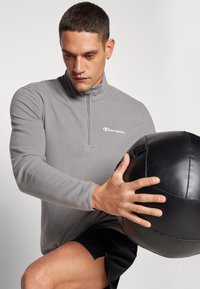 Sivi zip-up flís gornji dio s visokim ovratnikom, koji sadrži "Champion" logo u bijeloj boji. Model drži crnu medicinsku loptu.