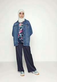 Casaco de ganga, blusa floral, calças largas azuis-marinho, sapatilhas cinzentas com detalhes laranja, e um hijab branco, apresentando um outfit em camadas.