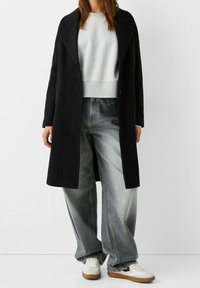 Svart lång kappa över en grå sweatshirt, kombinerad med överdimensionerade grå jeans och vita sneakers. Har en strukturerad silhuett och avslappnad passform.