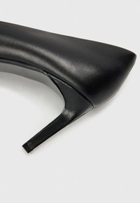 Zapato de tacón alto de cuero negro con punta afilada y tacón delgado. La superficie es suave con un ligero brillo y un detalle mínimo.