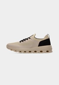 Chaussure de sport beige avec des accents noirs, dotée d'une tige texturée, d'une fermeture à lacets et d'une semelle légère et rembourrée avec des découpes circulaires.