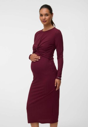 Femme enceinte aux cheveux foncés attachés en queue de cheval, portant une robe bordeaux à manches longues, tenant doucement son ventre, debout devant un fond uni.