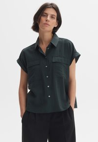 Blusa verde scuro a maniche corte con colletto strutturato, due tasche anteriori e chiusura con bottoni, caratterizzata da un tessuto testurizzato.