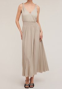 Robe longue beige sans manches avec un décolleté en V, des bretelles torsadées et une taille élastique ; plis fluides et finition en tissu texturé. Chaussures noires avec des accents de nœud.