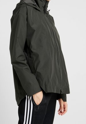Personne portant une veste à capuche zippée vert foncé et un pantalon noir à bandes blanches, main dans la poche, sur un fond clair uni.