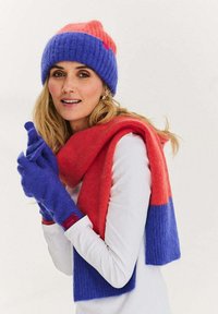 Zestaw czapka, szalik i rękawiczki na drutach. Czerwono-niebieskie pasy kolorów. Miękka tekstura, lekka. Szalik owinięty wokół szyi, rękawiczki na dłoniach.