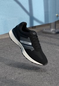 Black athletic shoe with a textured surface, featuring a white and gray stripe, a round toe, and a cushioned sole. Lightweight design.