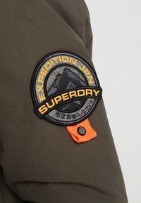 Ærme på en olivengrøn jakke med et rundt sort og gult mærke med teksten "EXPEDITION JPN" og "SUPERDRY," fremhævet af en orange løkke.