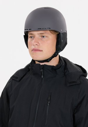 Grauer Skihelm mit Belüftungsöffnungen, verstellbarem Kinnriemen und weicher Ohrpolsterung. Getragen zu einer schwarzen Jacke mit Reißverschlusstasche.
