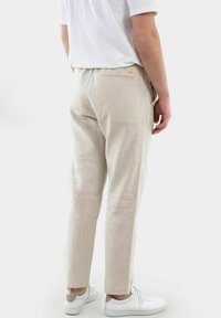 Helle beige Hosen aus einem strukturierten Stoff, mit einer Gesäßtasche mit kleinem Logo, die sich leicht zu den Knöcheln hin verjüngen.