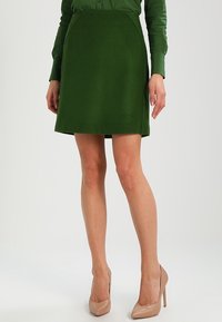 Falda verde de mezcla de lana en línea A con bolsillos laterales y textura suave, combinada con tacones altos beige.