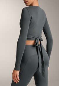 Dugih rukava siva bluzica s rebrastom teksturom i detaljem vezivanja na leđima, u kombinaciji s odgovarajućim rebrastim tajicama u istoj sivoj nijansi.