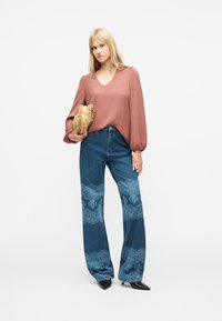 Γυναίκα που φοράει μια μπλούζα σε σκόνη ροζ και μπλε τζιν με ανοιχτό μπλε δαντέλα, κρατώντας μια μπεζ φάκελο με βολάν, στέκεται σε λευκό φόντο.