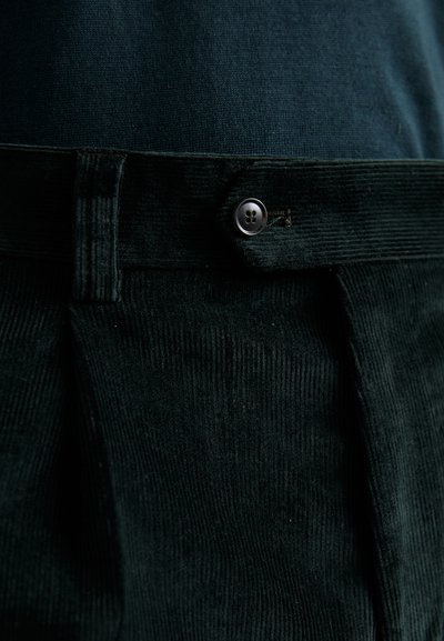 Close-up van donker groene corduroybroek met plooien, riemlus en knoopsluiting, gedragen met een donkerblauwe top.
