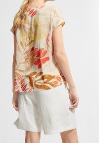 Bluse med blomsterprint i gennemsigtigt stof og korte ærmer, der har nuancer af orange, gul og beige samt en afslappet, løs pasform. Bæres med hvide shorts.