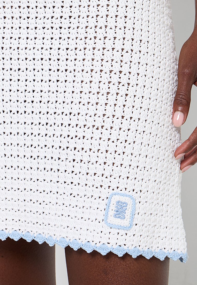 Weißes gehäkeltes Kleid mit strukturiertem Muster, gewelltem blauen Saum und kleinem blauen Markenlogo-Patch.