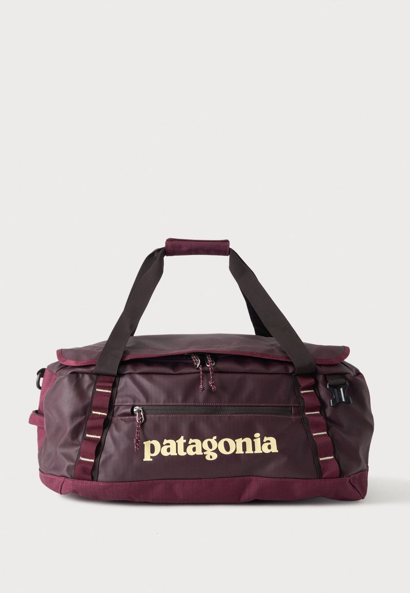 Σκούρο κόκκινη τσάντα Patagonia τύπου duffel με μαύρα λουριά και εμπρόσθια τσέπη με φερμουάρ σε απλό ανοιχτόχρωμο φόντο.