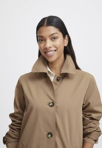 Cappotto trench brown con un ampio collo, dotato di chiusura a bottoni e tasche laterali. Tessuto liscio con una leggera finitura lucida.