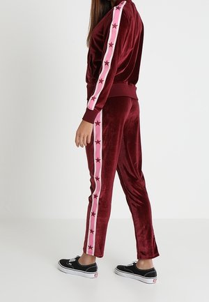 Kobieta w bordowym welurowym dresie z różową taśmą w gwiazdki na rękawach i nogawkach, w połączeniu z czarnymi trampkami.