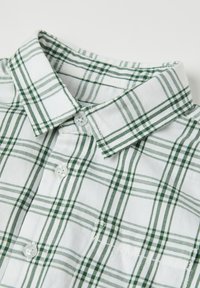 Napapaita valkoisessa ja vihreässä ruudullisessa kuosissa. Ominaisuuksia ovat klassinen kaulus, sileä puuvillakangas, kaksi nappia napituksessa ja taskuedusta.