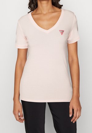 Kvinde iført en lyserød V-hals T-shirt med korte ærmer og et lille rødt Guess-logo på venstre bryst, kombineret med sorte bukser.