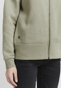 Olivovo zelená mikina na zips s rebrovanými manžetami a lemom, s malým čiernym logom na spodnej strane, spárovaná s čiernymi nohavicami.