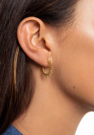 Goldene Creolen mit einem strukturierten Design und kleinen, hervorstehenden Perlen. Die Ohrringe sind kreisförmig und leicht, wodurch das Ohr betont wird.