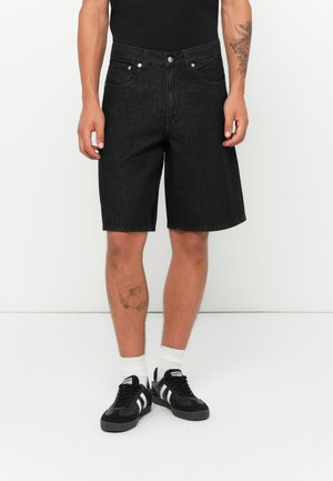 Man wearing black denim knee-length shorts, black sneakers with white stripes, white socks, and visible tattoos on left forearm and lower right leg.