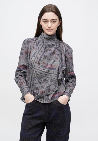 Szürke blúz virágmintákkal, magas fodros gallérral és hosszú ujjakkal. Sötét, strukturált nadrággal párosítva.