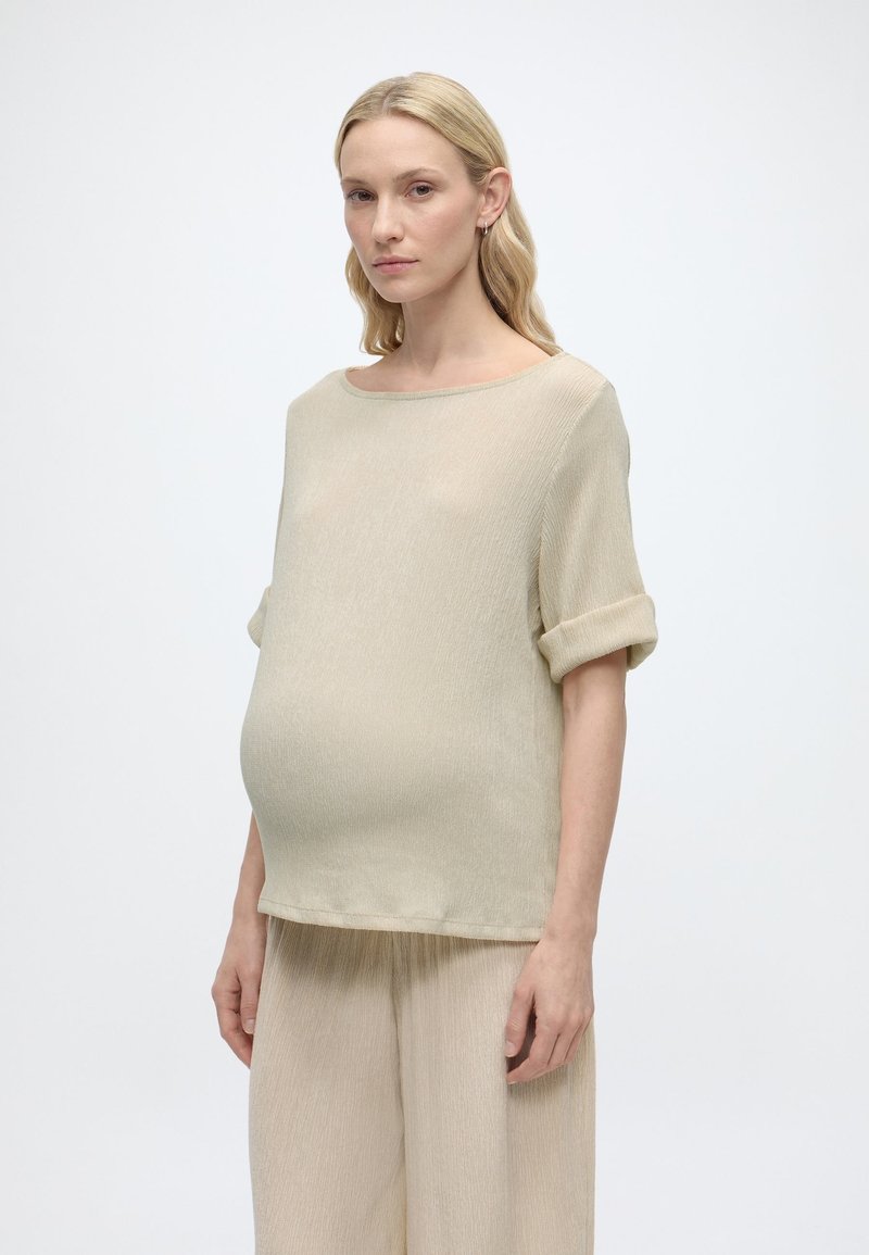 Έγκυος γυναίκα με μακριά ξανθά μαλλιά, ντυμένη με μπεζ υφασμάτινο χαλαρό τοπ και παντελόνι, στέκεται μπροστά σε λευκό φόντο.