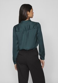 Blusa in satin di un blu scuro con cucitura a yoke sul retro, pieghe e maniche lunghe, abbinata a pantaloni neri con vestibilità rilassata e vita elastica.