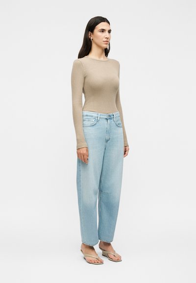 Γυναίκα όρθια, φοράει μπεζ ριμπ πλεκτό μακρυμάνικο τοπ, ανοιχτό μπλε παντελόνι με φαρδιά μπατζάκια και μπεζ σανδάλια με τακούνι σε λευκό φόντο.