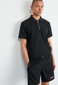 Melns polo krekls ar rāvējslēdzēja apkakli, teksturētu audumu un mazu logo. Saskaņots ar melniem šortiem, kuru priekšpusē ir rozā logo un kabatas.