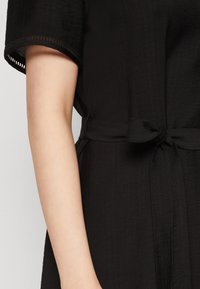 Käsi ja torso, kanda musta tekstuuriga lühikeste varrukatega rõivastust, mille vöökoht on seotud kangast vöödiga.