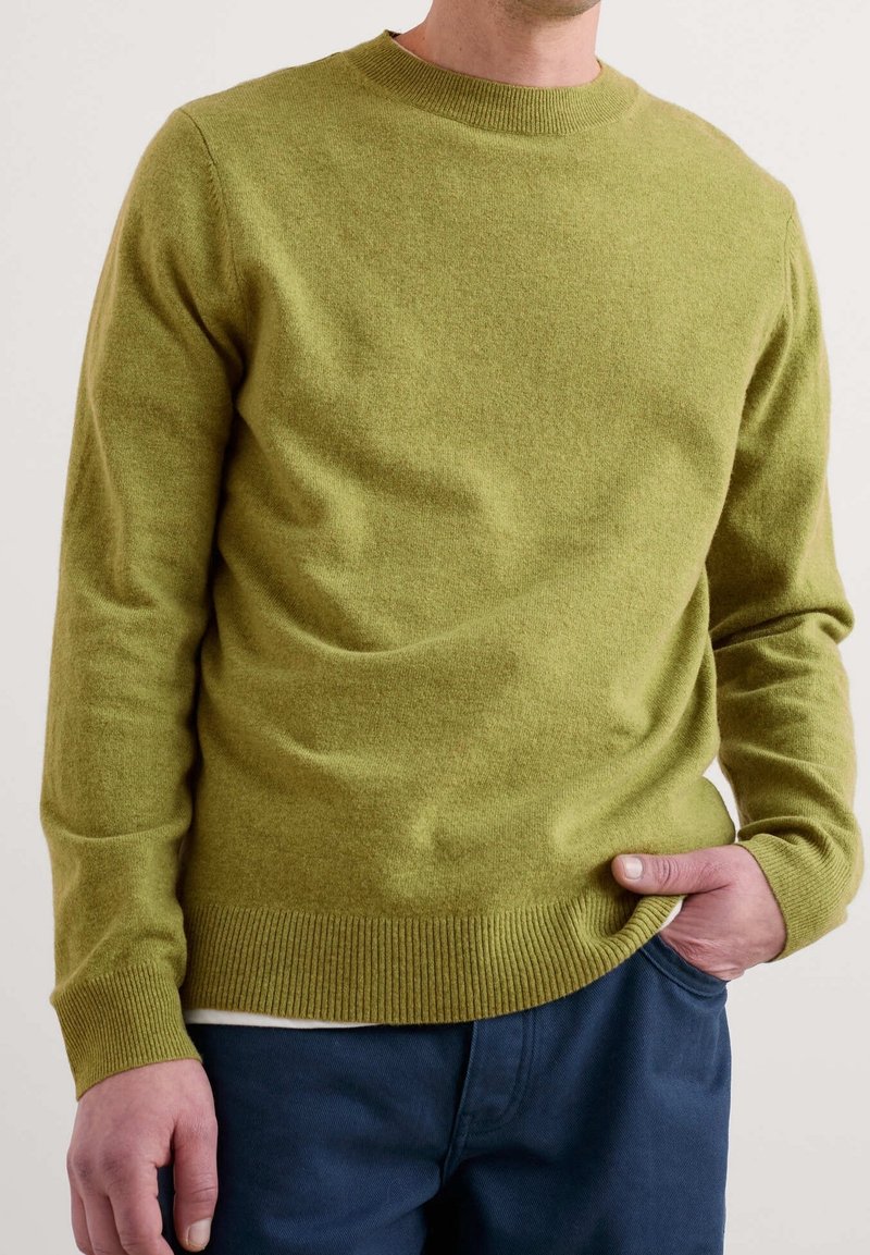 Homme portant un pull ras du cou vert olive et un pantalon bleu marine, avec une main dans la poche, sur un fond uni.