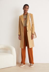 Manteau en laine beige porté sur un pull côtelé olive et un pantalon couleur rouille, associé à des chaussures marron clair avec une tige texturée. Sac à bandoulière simple.