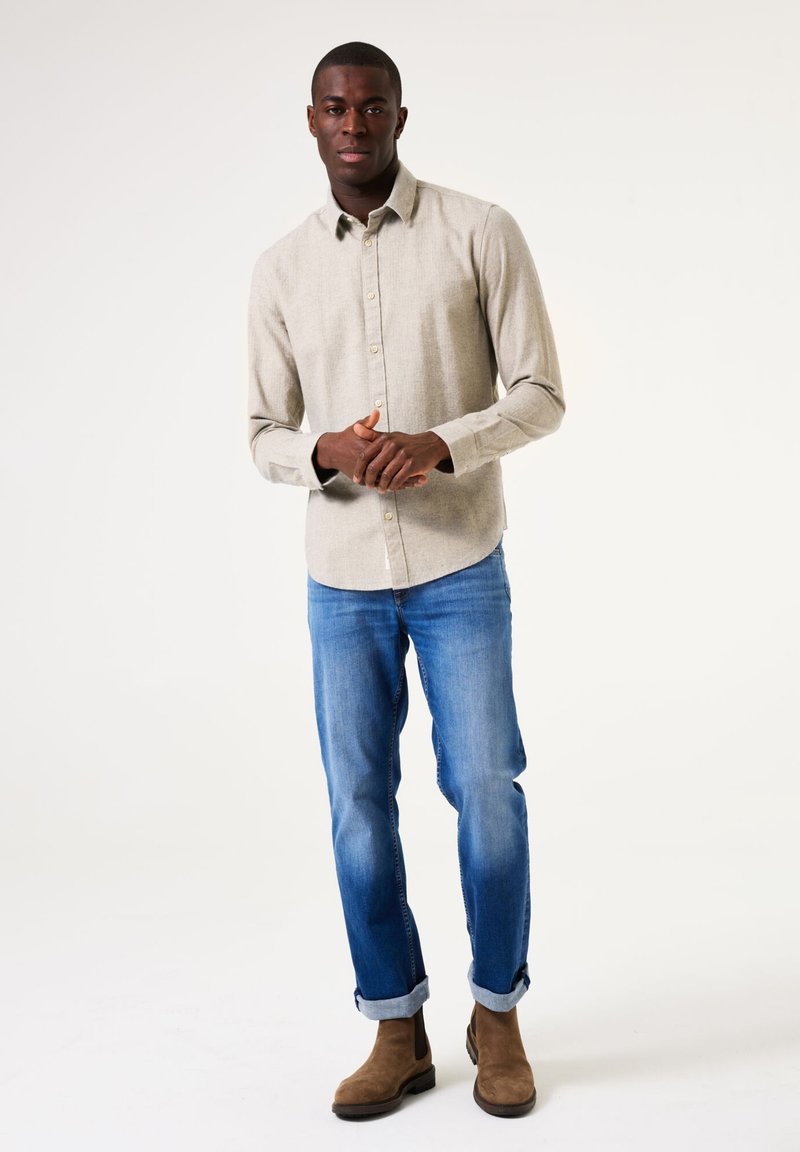 Beige, button-down shirt van een structuurstof, gedragen met lichtblauwe jeans en bruine Chelsea boots, opgerold aan de mouwen.