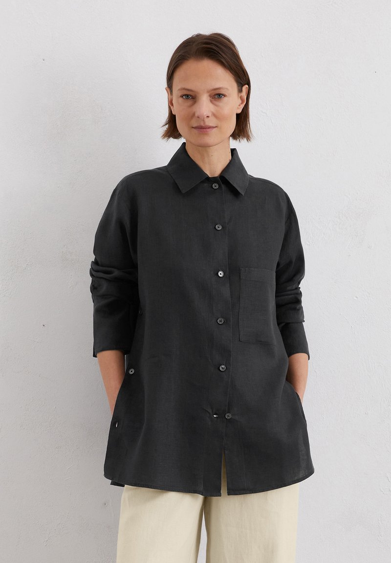 Schwarze Leinenbluse mit Kragen, einer einzelnen Brusttasche und durchgehender Knopfleiste. Verfügt über lange Ärmel mit umgeschlagenen Bündchen und seitlichen Schlitzen.