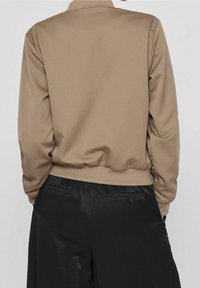 Personne portant une veste beige à manches longues avec poignets et ourlet élastiques, associée à un pantalon large en satin noir, vue de dos.