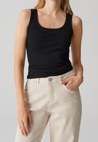 Femme portant un haut noir sans manches à encolure dégagée et un pantalon beige taille haute, avec une main dans la poche et un fond neutre.